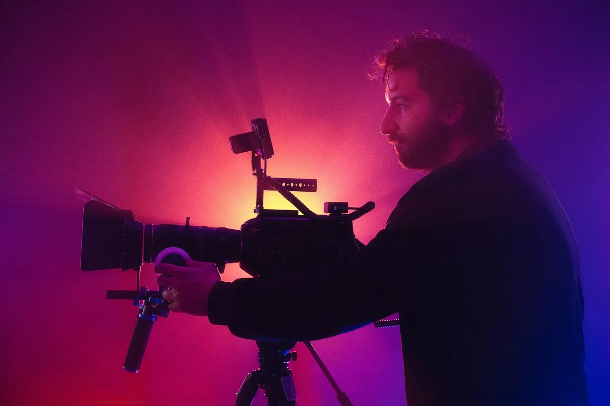 Camera operator in silhouette behind a cinema camera, lit by pink and purple studio light
