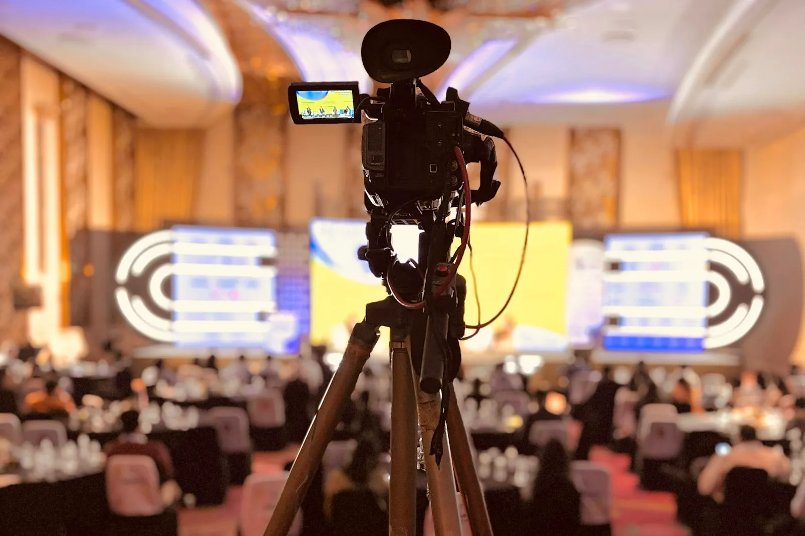 Video camera on a tripod at the back of a hotel ballroom, filming a conference stage with large screens and a seated audience