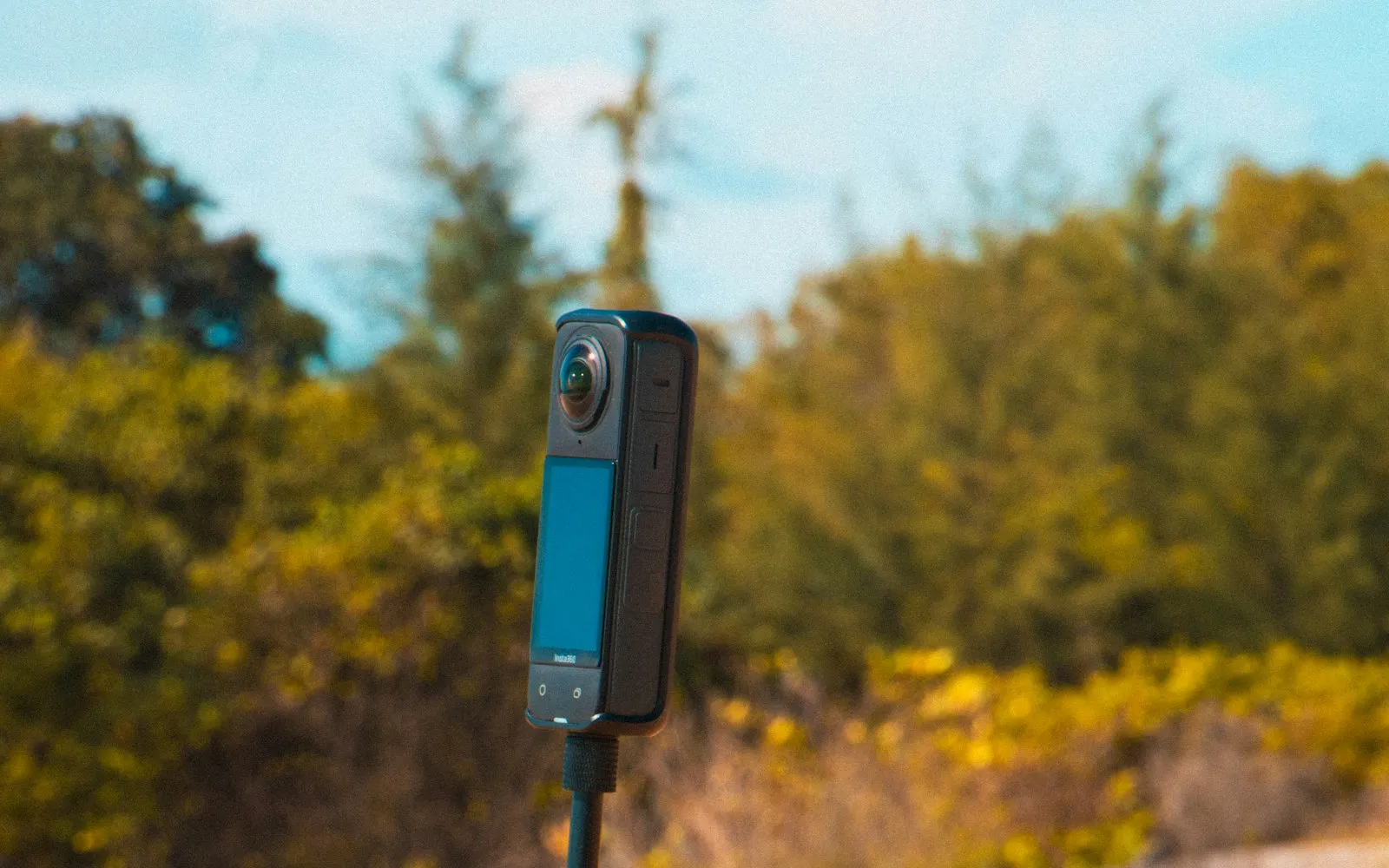 A compact 360-degree camera on a selfie stick planted in the sand at a beach, with trees behind