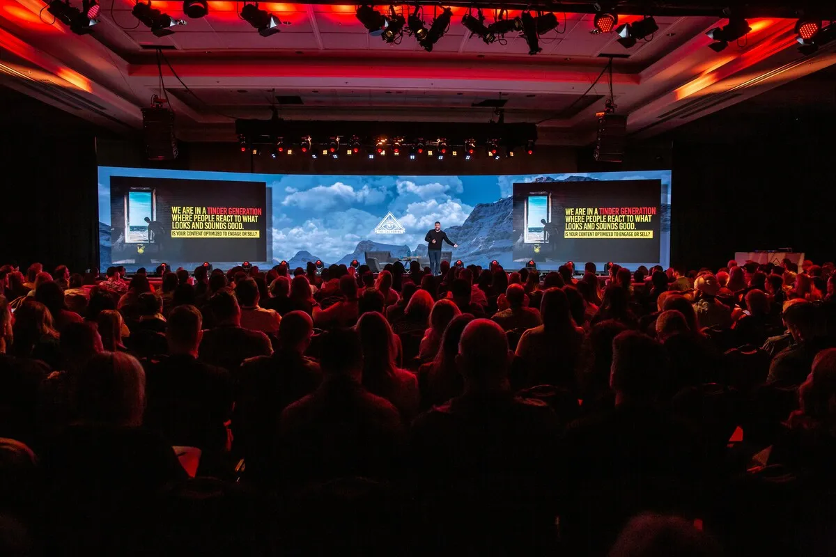 Speaker on a conference stage in front of a wide LED screen, seen from behind a packed audience
