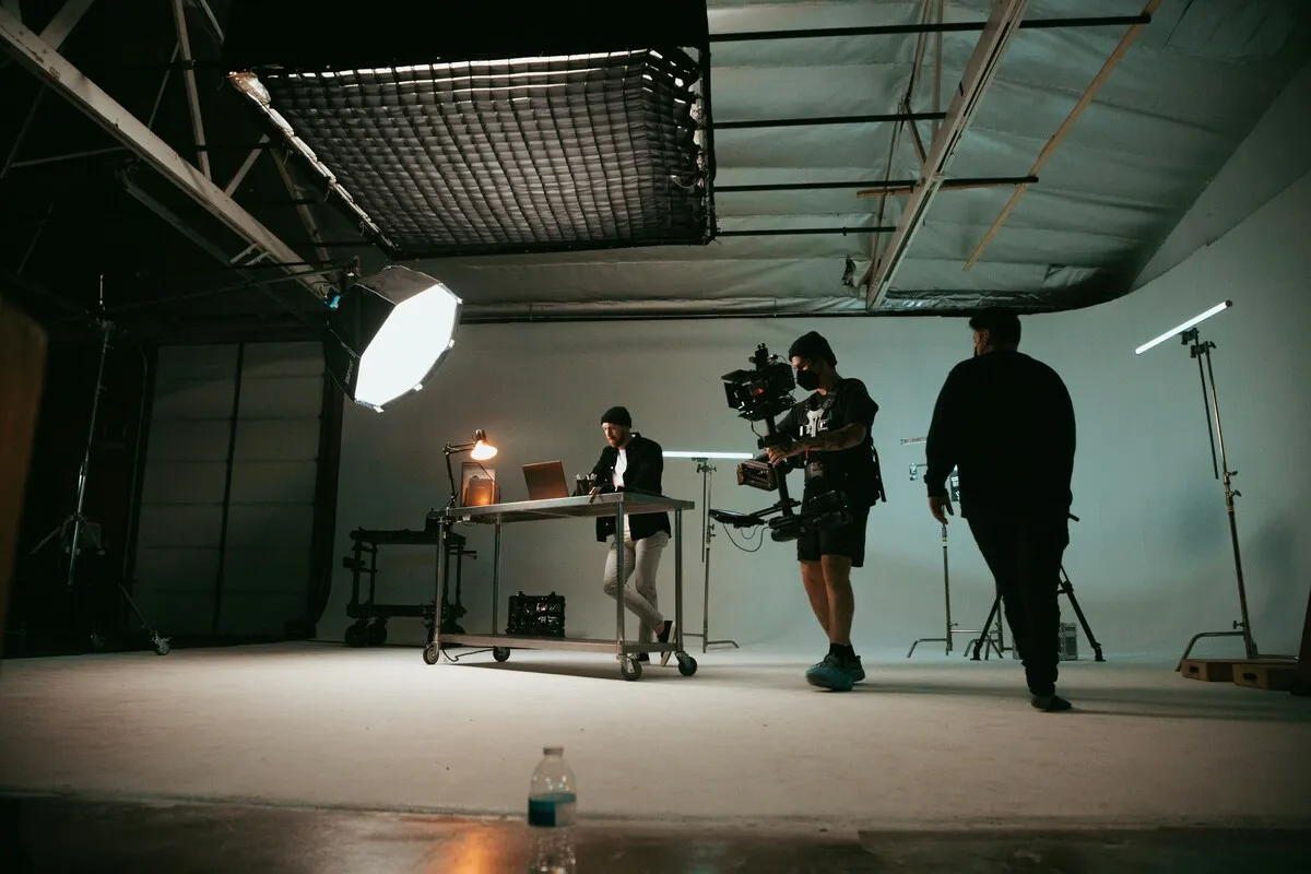 Camera operator with a gimbal rig filming a scene on a studio set, with large softbox lights overhead