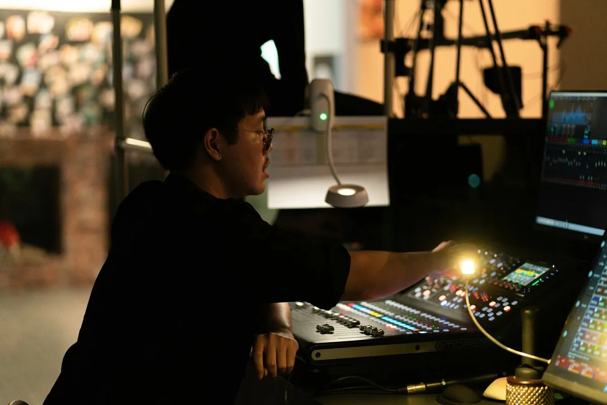 Sound engineer leaning over a digital mixing desk in a dim control area, with level meters on a screen beside him