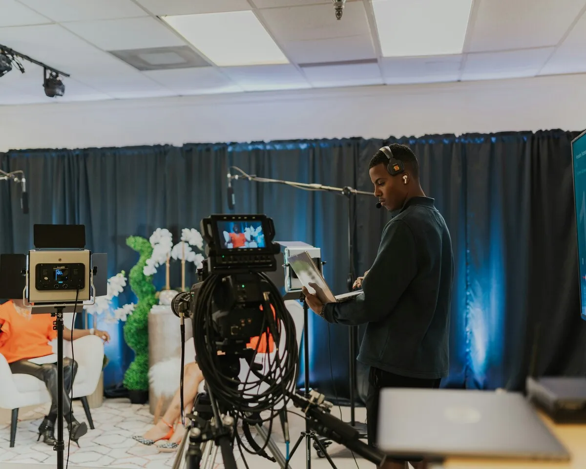 Camera operator with a headset and clipboard beside a studio camera filming a two-person interview set in front of blue-lit curtains