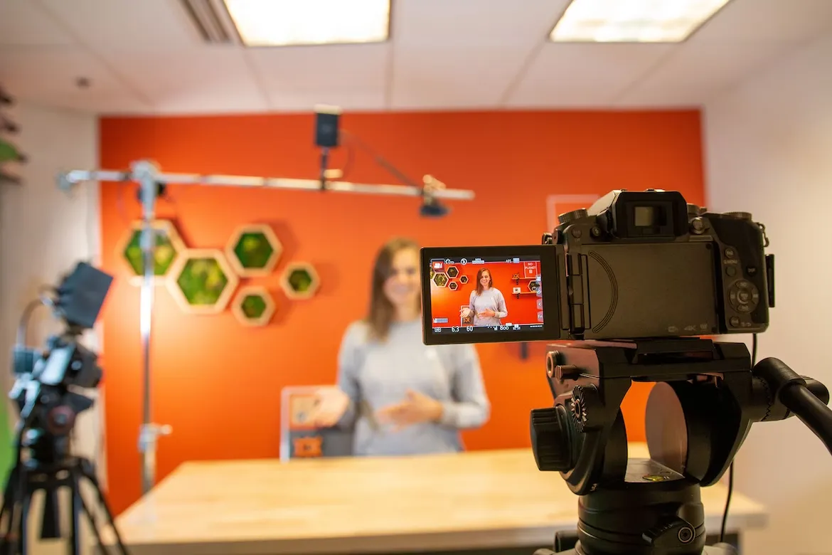 A presenter filmed in front of an orange wall, seen on the flip-out screen of a camera on a tripod in the foreground